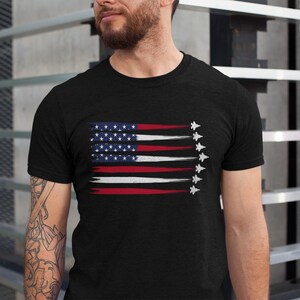 Puede incluir: Una camiseta negra con un dise&ntilde;o de bandera estadounidense desgastado y una fila de aviones de combate blancos volando en formaci&oacute;n.
