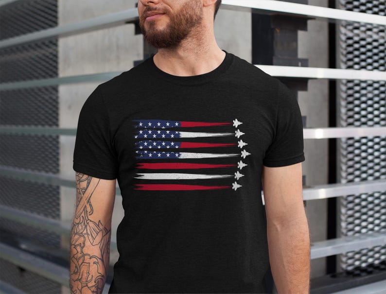 Puede incluir: Una camiseta negra con un dise&ntilde;o de bandera estadounidense desgastado y una fila de aviones de combate blancos volando en formaci&oacute;n.