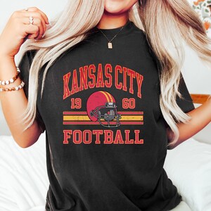 K&ouml;nnte beinhalten: Schwarzes T-Shirt mit einem Grafikdruck im Vintage-Stil eines Footballhelms und dem Text "Kansas City Football 19 60".