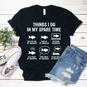 May include: Black t-shirt with white text that reads "Things I do in my spare time". The text is arranged in a grid with six icons of fish and aquariums. The icons include "Buy fish for my aquarium", "Look at my aquarium", "Research aquariums", "Talk about my aquarium", "Think about my aquarium", and "Dream about my aquarium".