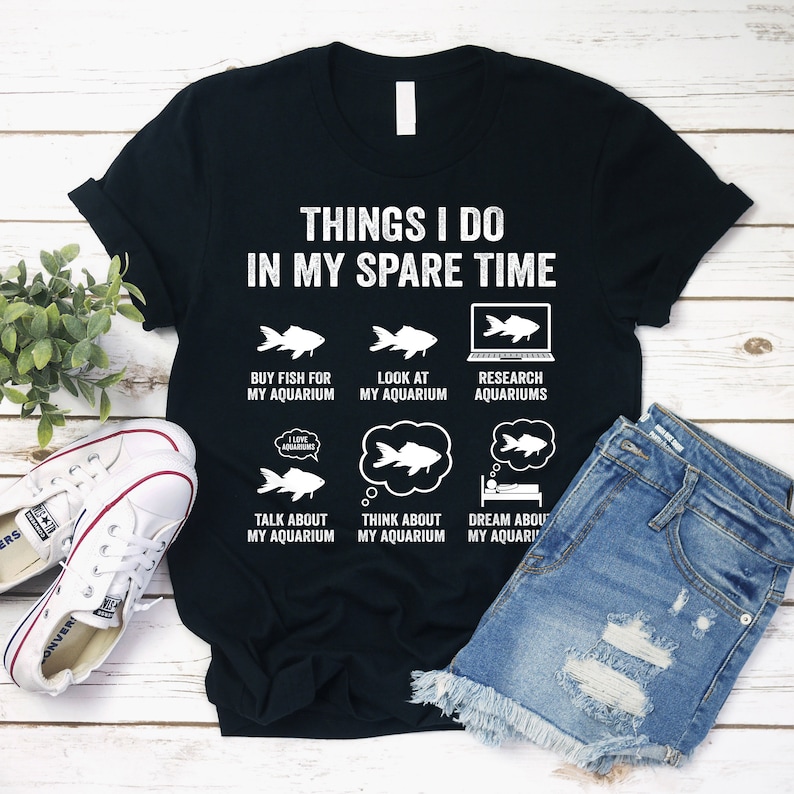 May include: Black t-shirt with white text that reads "Things I do in my spare time". The text is arranged in a grid with six icons of fish and aquariums. The icons include "Buy fish for my aquarium", "Look at my aquarium", "Research aquariums", "Talk about my aquarium", "Think about my aquarium", and "Dream about my aquarium".