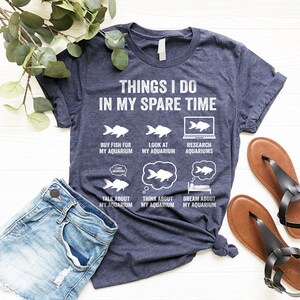 May include: A navy blue t-shirt with white text that reads "Things I do in my spare time". The text is surrounded by illustrations of fish and aquariums. The illustrations include the text "Buy fish for my aquarium", "Look at my aquarium", "Research aquariums", "Talk about my aquarium", "Think about my aquarium", and "Dream about my aquarium".