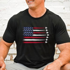 Puede incluir: Camiseta negra con un dise&ntilde;o de bandera estadounidense desgastado y una fila de aviones de combate volando en formaci&oacute;n.