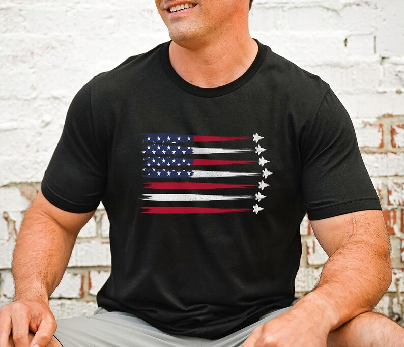 Puede incluir: Camiseta negra con un dise&ntilde;o de bandera estadounidense desgastado y una fila de aviones de combate volando en formaci&oacute;n.