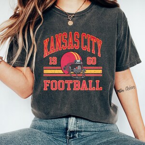 K&ouml;nnte beinhalten: Ein dunkelgraues T-Shirt mit einer Grafik im Vintage-Stil eines Footballhelms und dem Text "KANSAS CITY 19 60 FOOTBALL".