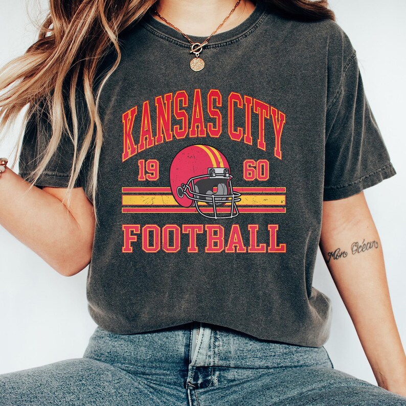 K&ouml;nnte beinhalten: Ein dunkelgraues T-Shirt mit einer Grafik im Vintage-Stil eines Footballhelms und dem Text "KANSAS CITY 19 60 FOOTBALL".