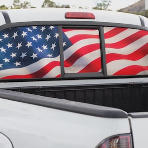May include: White pickup truck with a rear window featuring a vibrant American flag design. The flag's stars and stripes are prominently displayed across the window's three panels, creating a patriotic statement.