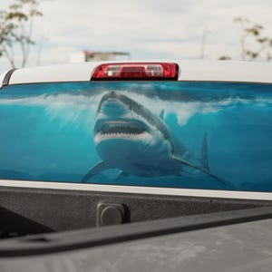 May include: A white truck's rear window features a large, detailed image of a great white shark swimming in blue water. The shark is facing forward with its mouth open, teeth visible. The background shows sunlight filtering through the water.