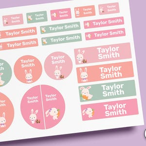 Puede incluir: Una hoja de etiquetas adhesivas de varias formas y tamaños, con una ilustración de un conejo de dibujos animados y el nombre impreso Taylor Smith. Las pegatinas son de tonos rosa y verde salvia, con el texto STICK-ON visible en el lateral.