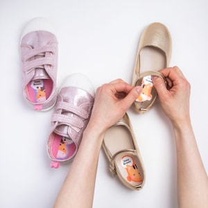 May include: Two pairs of shoes, one pair of metallic pink trainers with hook-and-loop straps and one pair of gold ballet flats with a strap. Inside each shoe is a custom adhesive label featuring a cartoon animal graphic and the text Joey Brown.