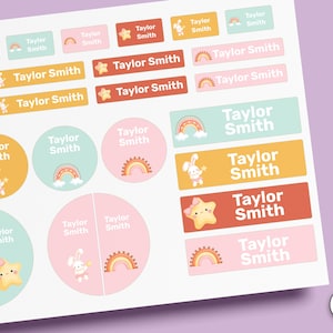 May include: A sheet of assorted rectangular and circular stick-on labels in pastel colours featuring the name Taylor Smith. The designs include illustrations of rainbows, stars, and bunnies. The text STICK-ON is visible on the right side.
