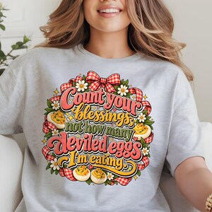 Peut inclure: Un t-shirt gris clair avec un motif coloré portant l'inscription "Count your blessings, not how many deviled eggs I'm eating." Le design comprend des rubans à carreaux rouges et blancs, des feuilles vertes, des marguerites et des œufs farcis.
