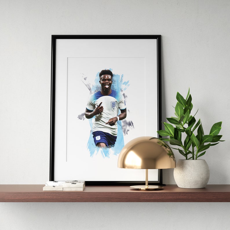 Puede incluir: Una impresi&oacute;n enmarcada de un jugador de f&uacute;tbol con una camiseta blanca y azul sobre un fondo de acuarela. El jugador sonr&iacute;e y levanta la mano derecha en el aire. El marco es negro y la impresi&oacute;n est&aacute; sobre un fondo blanco.