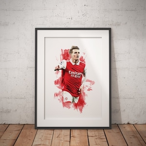 May include: A framed print of a soccer player wearing a red and white jersey with the text "Emirates Fly Better" on the front. The player is running with the ball and is surrounded by a red and white watercolor background.