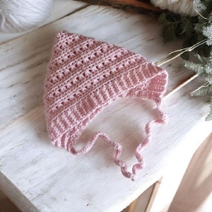 Pode incluir: Um gorro de bebé de crochê rosa com uma longa gravata. O gorro é feito com um padrão de ponto delicado e tem um visual suave e feminino.