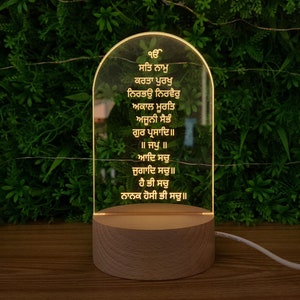 Puede incluir: Una luz nocturna LED brillante con una base de madera. El panel acr&iacute;lico transparente muestra texto grabado en formato vertical. La luz emite un brillo c&aacute;lido e invitador, y la base es de color madera natural. Un cable de alimentaci&oacute;n blanco sale de la base.