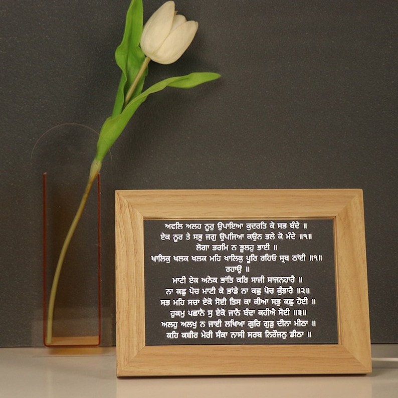 Puede incluir: Un marco de madera con fondo negro y texto blanco en punjabi. El texto es un poema sobre la naturaleza de Dios y la importancia de la verdad. El marco est&aacute; sobre una superficie gris con un tulip&aacute;n blanco en un jarr&oacute;n de vidrio.