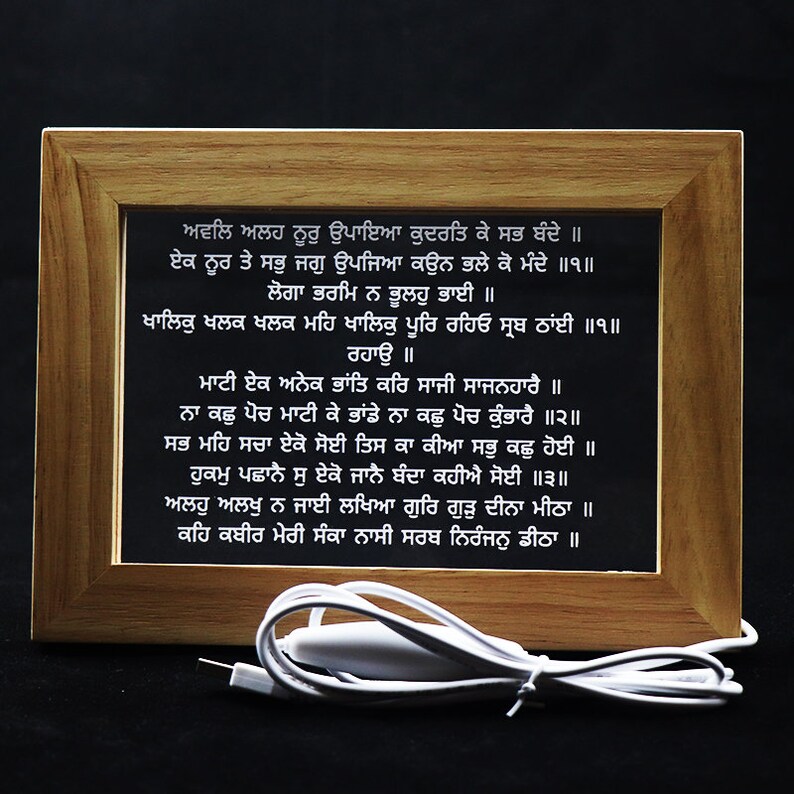 Puede incluir: Una impresi&oacute;n enmarcada con un fondo blanco y texto negro en idioma punjabi. El texto es una cita religiosa. El marco es de madera y tiene un acabado marr&oacute;n claro. La impresi&oacute;n est&aacute; iluminada por un cable blanco.