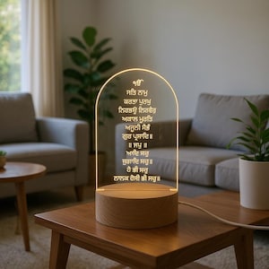 Puede incluir: L&aacute;mpara LED con base de madera y panel de acr&iacute;lico transparente arqueado. El panel muestra texto dorado e iluminado con un brillo c&aacute;lido. La l&aacute;mpara est&aacute; sobre una mesa de madera.