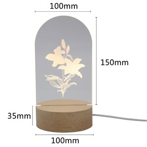 Puede incluir: L&aacute;mpara LED decorativa con dise&ntilde;o floral. Tiene un panel acr&iacute;lico transparente arqueado con una flor de lirio grabada, de 15 cm de alto y 10 cm de ancho. Se apoya en una base de madera de 10 cm de ancho y 3,5 cm de alto.