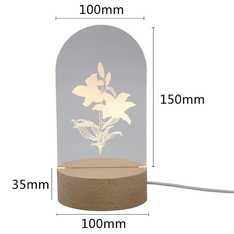 Puede incluir: L&aacute;mpara LED decorativa con dise&ntilde;o floral. Tiene un panel acr&iacute;lico transparente arqueado con una flor de lirio grabada, de 15 cm de alto y 10 cm de ancho. Se apoya en una base de madera de 10 cm de ancho y 3,5 cm de alto.