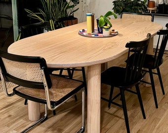 May include: A light wood oval dining table with four black chairs. The table has a natural wood finish and thick legs. Two of the chairs have woven cane seats and backs. The table is set with a colorful tray and two small decorative bottles.