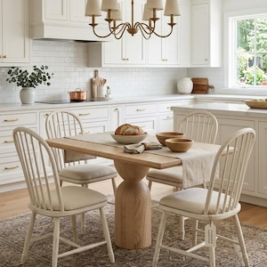 May include: A rectangular light wood dining table with a thick cylindrical pedestal base, surrounded by four off-white spindle-back chairs with upholstered seats. The table is set with a beige runner, two wooden bowls, and a loaf of bread.