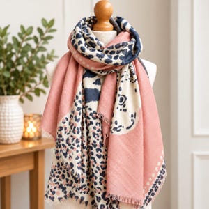 May include: A soft scarf featuring a dual-pattern design with a dusty pink solid section and a cream-coloured section adorned with a dark navy blue leopard print. The scarf has a frayed edge finish and is draped around a mannequin neck form.