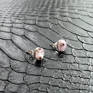 Pode incluir: Um par de brincos de pressão de prata com detalhes em rosa e branco. Os brincos estão sobre uma superfície preta texturizada.