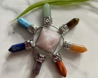 May include: A chakra crystal healing set with seven chakra stones in a silver metal frame. The stones are arranged in a circle around a pink rose quartz crystal. The stones are green, blue, turquoise, indigo, purple, orange, and yellow.