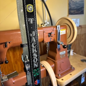 May include: Black leather belt with the name "T JACKSON" in silver letters. The belt has a gray suede lining and decorative pins, including a clover and an emergency medical services symbol. A sewing machine is in the background.