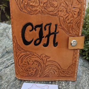 May include: A tan leather journal with tooled floral designs and the initials "C.J.H." embossed in black. The journal has a brown leather strap with a metal button closure.