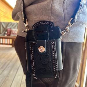 May include: Black leather crossbody bag with a floral embossed design and pink stitching. The bag has a flap closure with a silver button and a black leather strap. The bag is worn over the shoulder.