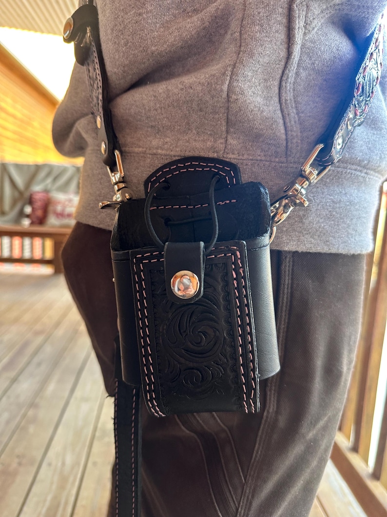 May include: Black leather crossbody bag with a floral embossed design and pink stitching. The bag has a flap closure with a silver button and a black leather strap. The bag is worn over the shoulder.