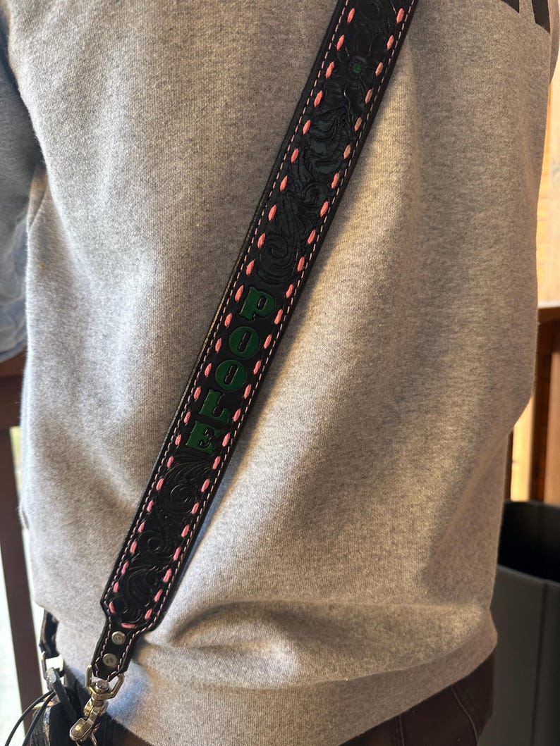 May include: Black leather bag strap with embossed floral design and pink stitching. The word "POOLE" is displayed in green lettering. The strap is attached to a bag with a silver clasp.