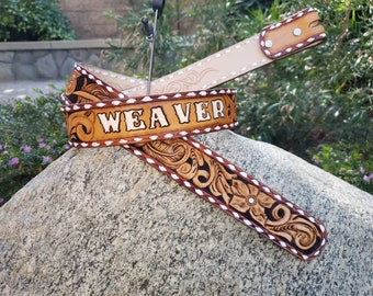May include: A brown leather belt with intricate floral carving and a white stitched border. The belt is personalized with the name "WEAVER" in white lettering.