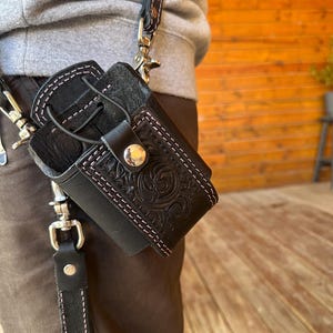 May include: Black leather holster with pink stitching and a decorative embossed design. The holster is attached to a black strap with silver-tone hardware and a patterned strap. The holster is designed to hold a small item.