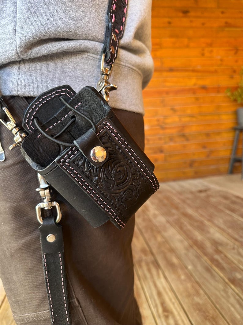 May include: Black leather holster with pink stitching and a decorative embossed design. The holster is attached to a black strap with silver-tone hardware and a patterned strap. The holster is designed to hold a small item.