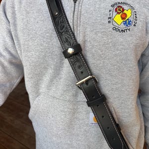 May include: Black leather shoulder strap with floral tooling and white stitching. The strap has a silver buckle and a silver button closure. The top edge has pink stitching. The strap is worn over a gray hoodie.