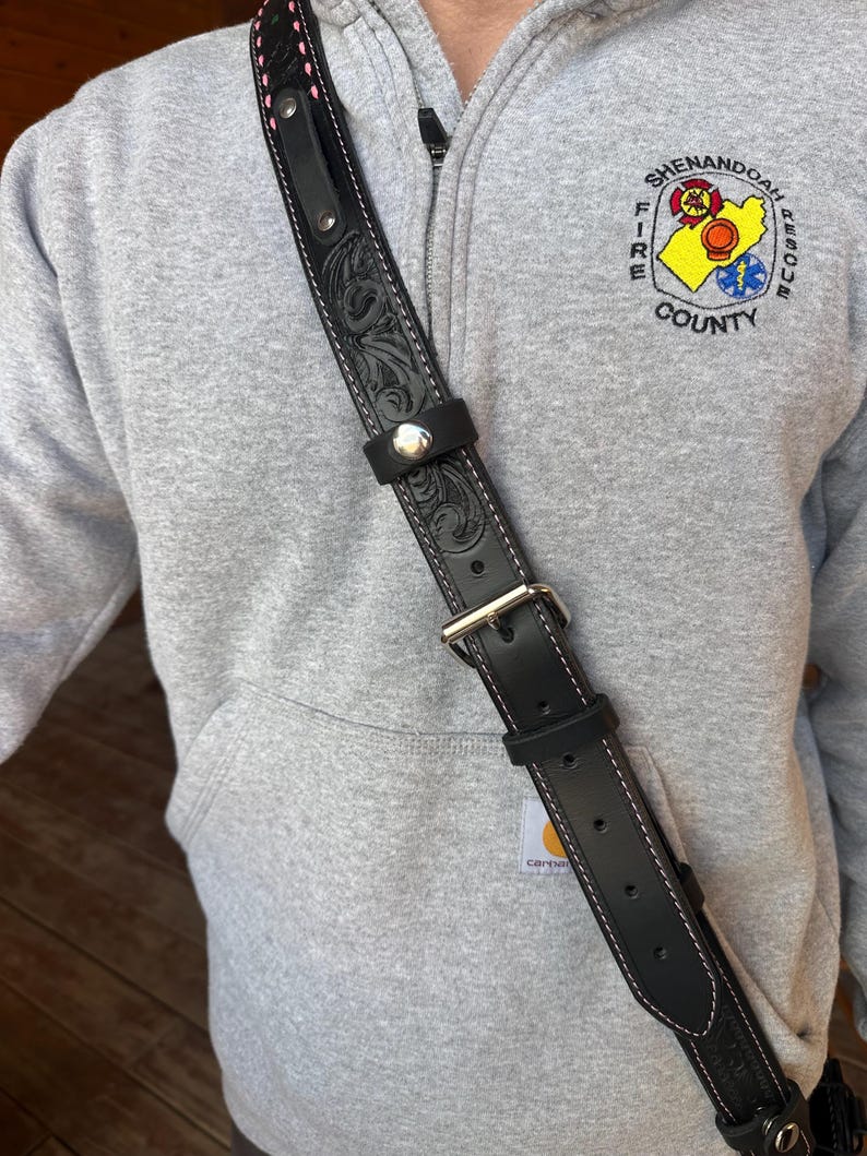 May include: Black leather shoulder strap with floral tooling and white stitching. The strap has a silver buckle and a silver button closure. The top edge has pink stitching. The strap is worn over a gray hoodie.