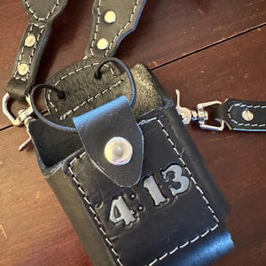 May include: Black leather radio case with white stitching and a shoulder strap. The case has the text "4:13" embossed on the front. The strap and case are connected with metal hardware.