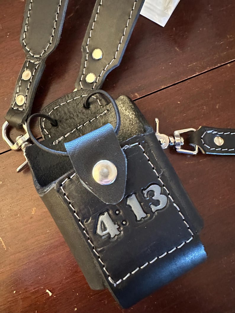 May include: Black leather radio case with white stitching and a shoulder strap. The case has the text "4:13" embossed on the front. The strap and case are connected with metal hardware.