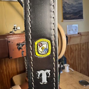 May include: Close-up of a dark brown leather belt with white stitching along the edges. The belt features a metal emblem with the words "Shenandoah County" and the letter "T" in silver. The belt is likely handmade.