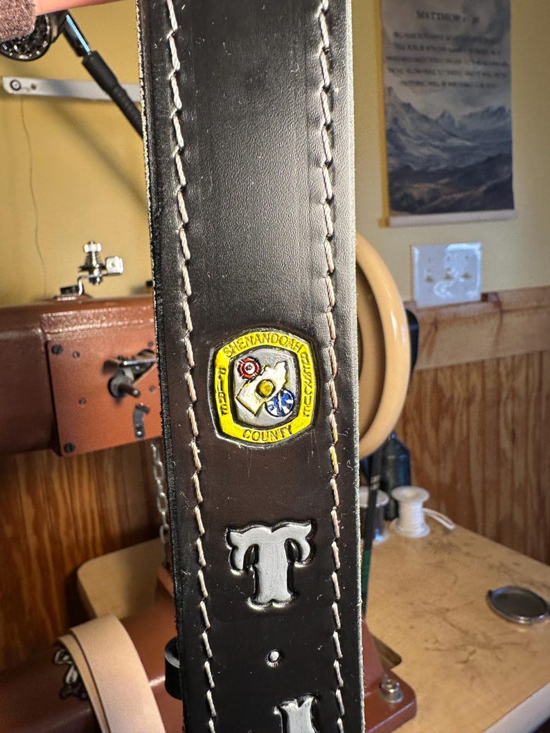 May include: Close-up of a dark brown leather belt with white stitching along the edges. The belt features a metal emblem with the words "Shenandoah County" and the letter "T" in silver. The belt is likely handmade.
