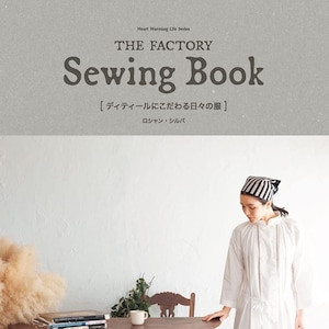 Könnte beinhalten: Ein weißes Nähbuch mit dem Titel "The Factory Sewing Book" und dem Untertitel "[Detail zu Alltagskleidung]" in Japanisch. Der Buchumschlag zeigt eine Frau in einem weißen Kleid und einem schwarz-weiß gestreiften Stirnband. Das Buch liegt auf einem Holztisch mit einer Tasse und einem Stapel Bücher.