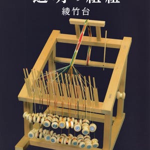 May include: A light-colored wooden Ayatakedai braiding stand with multiple vertical pegs and weighted bobbins holding colorful threads. The threads converge into a braided strip at the top. Text reads: 道明の組紐 綾竹台 and 道明古式糸組法教処.