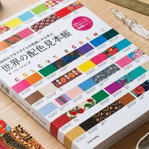 以下が含まれることがあります： 「COLOUR SCHEME」というタイトルのデザインブック。白い表紙にはカラフルなテキスタイルパターンの列と日本語のテキストが印刷されています。本は木製のテーブルの上に置かれ、陶器製の絵具パレット、ガラスペン、布のサンプルが添えられています。