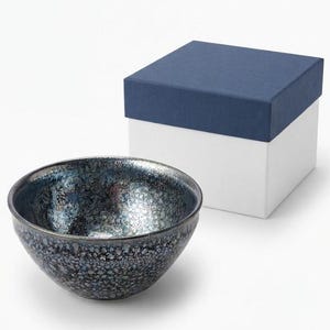 May include: A small, round ceramic bowl with a dark, iridescent, speckled glaze featuring metallic blue and silver tones. The bowl is accompanied by a square, two-piece gift box with a white base and a solid dark blue lid, presented together.