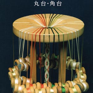 May include: A wooden marudai braiding stand with a circular top and vertical legs. Multiple colored threads extend from the center through slots to wooden bobbins hanging in a ring. The text reads: 道明の組紐 丸台・角台 and 道明古式糸組法教処.