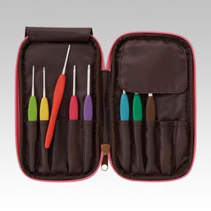 Puede incluir: Un juego de ocho ganchillos con mangos de colores guardados en un estuche de tela marrón con cremallera rosa. Los ganchillos tienen puntas metálicas y mangos ergonómicos en verde, amarillo, rojo, rosa, morado, azul claro, verde oscuro y marrón.